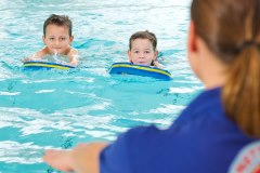Zwei Kinder mit Schwimmbrettern im Wasser und eine lehrende Person